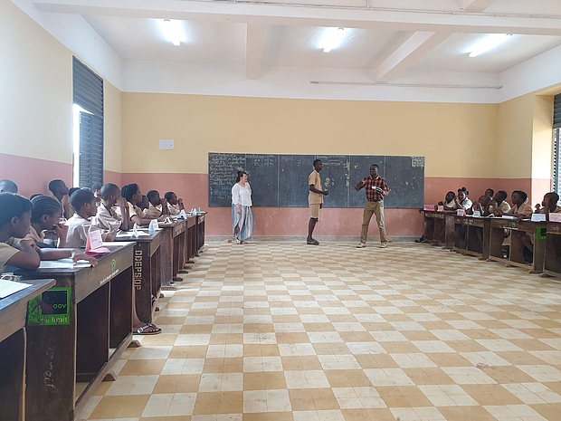Foto aus einem Klassenraum in Porto Novo. An der Tafel stehen drei Lehrende. LInks und rechts an den Wänden sitzen Schulkinder und folgen dem Unterricht.
